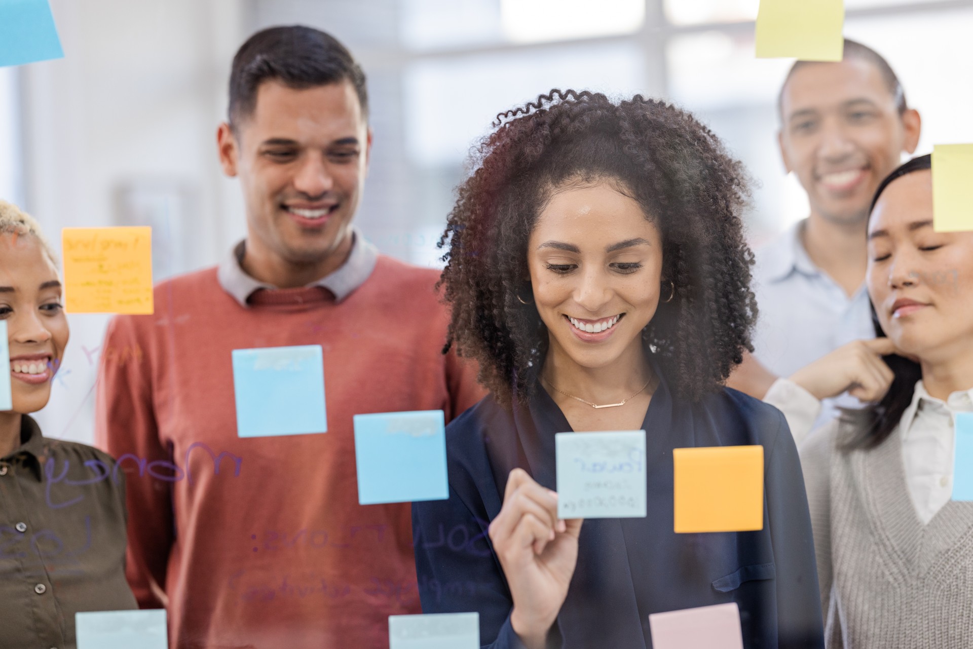 Black woman, writing and schedule planning with team in strategy or brainstorming on glass wall at office. African American female designer in teamwork project plan, tasks or post it with sticky note